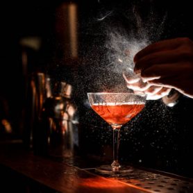 Barmans hands sprinkling the juice into the cocktail glass filled with alcoholic drink on the dark background